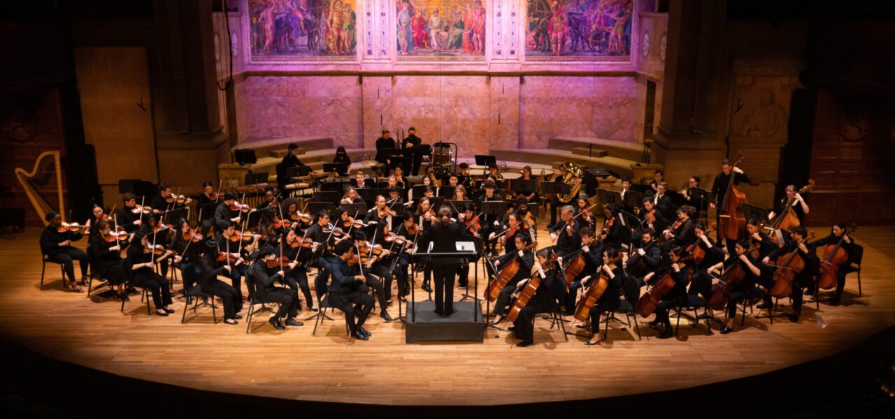 Many Sinfonia ensemble performers seated on a large stage, with their instruments