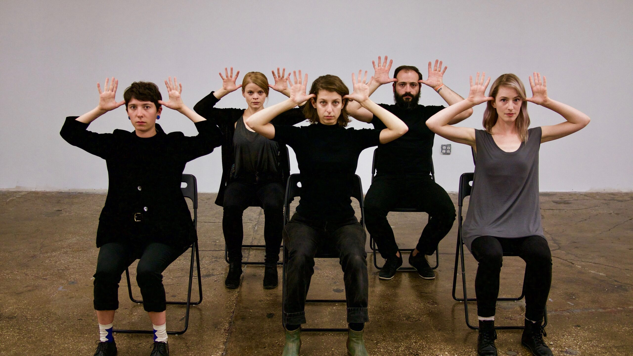A group of people wearing black ensemble and sitting on chairs with their hands up